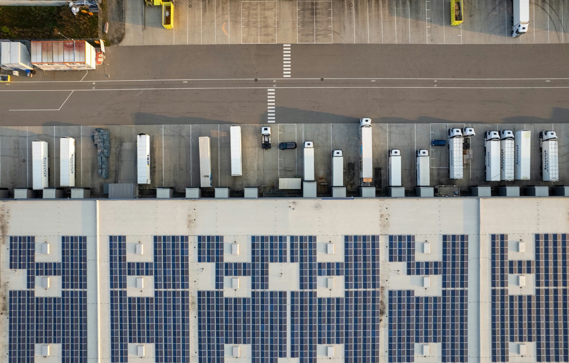 Vista aérea de galpão logístico com telhado fotovoltaico, docas de carga e frota de carretas em pátio pavimentado.